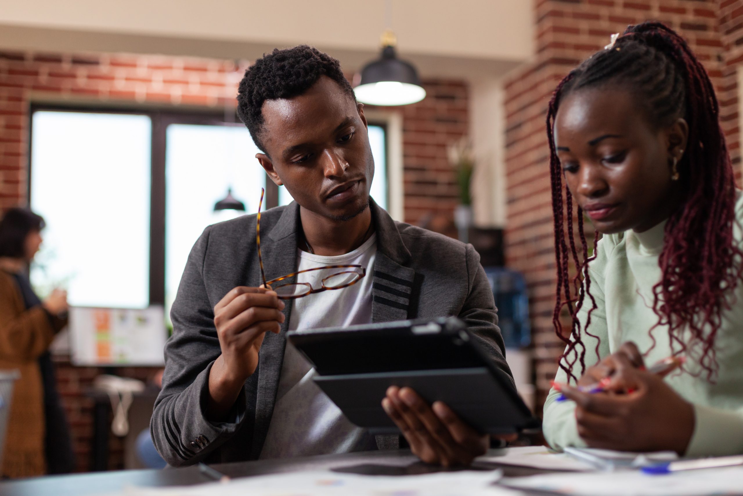 Two young professionals discussing financial planning strategies, one holding a tablet and the other taking notes, in a modern office setting.